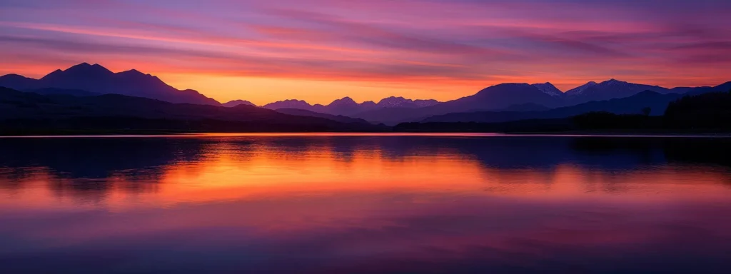 a breathtaking sunset casts vibrant hues of orange and purple across a tranquil lake, reflecting the silhouette of majestic mountains in the distance.