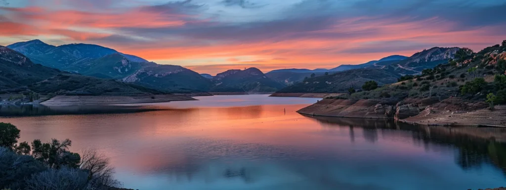 a serene landscape unfolds at sunrise, with a tranquil lake reflecting vibrant hues of orange and pink, framed by majestic mountains under a soft, glowing sky.